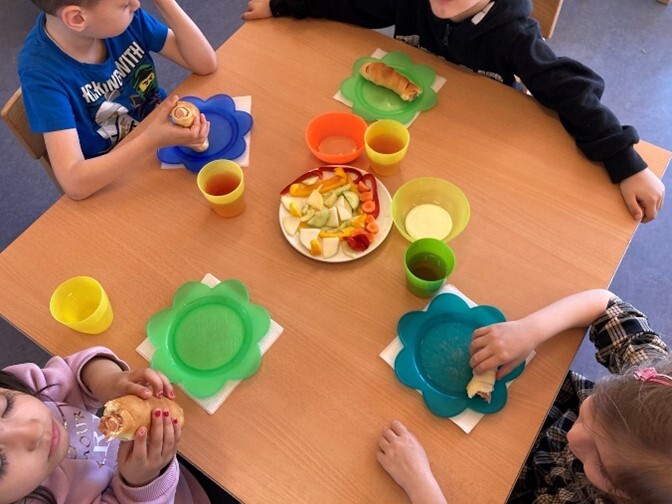 Vier Kinder sitzen zu Tisch und essen aus Plastikgeschirr Salat und eine Wirscecke